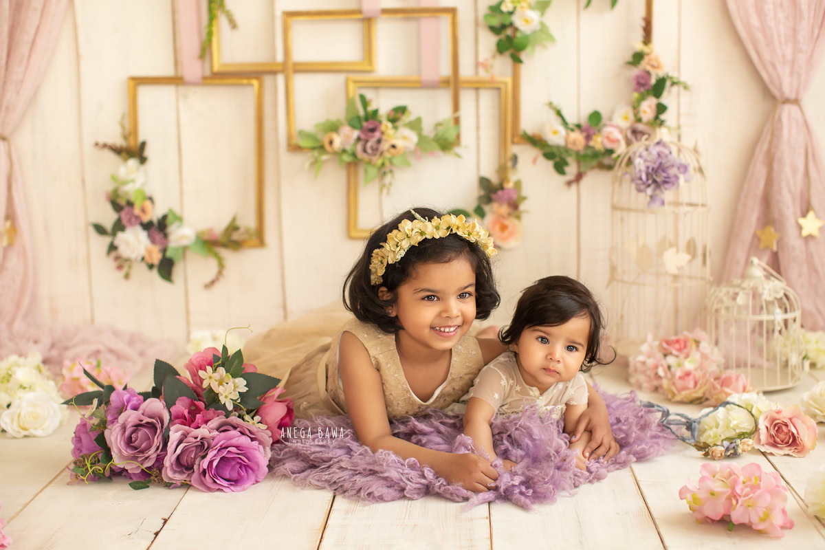 In a delightful photoshoot at Anega Bawa Photography's studio in Delhi and Gurgaon, your 14-month to 1-year-old girl and her sibling are captured in a serene scene. Seated on a lavender rug against a soothing beige backdrop, surrounded by lavender flowers, charming castles, and a floral frame on the wall, they create enchanting memories of their sibling bond.