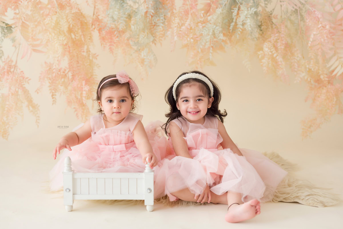 Celebrate the precious bond between siblings with Anega Bawa Photography in Delhi and Gurgaon. Your 14-month to 1-year-old girl, accompanied by her sibling, is captured against a charming leafy peach backdrop, wearing a cute headband that adds to her adorable charm. Book your session today to immortalize these special moments forever.
