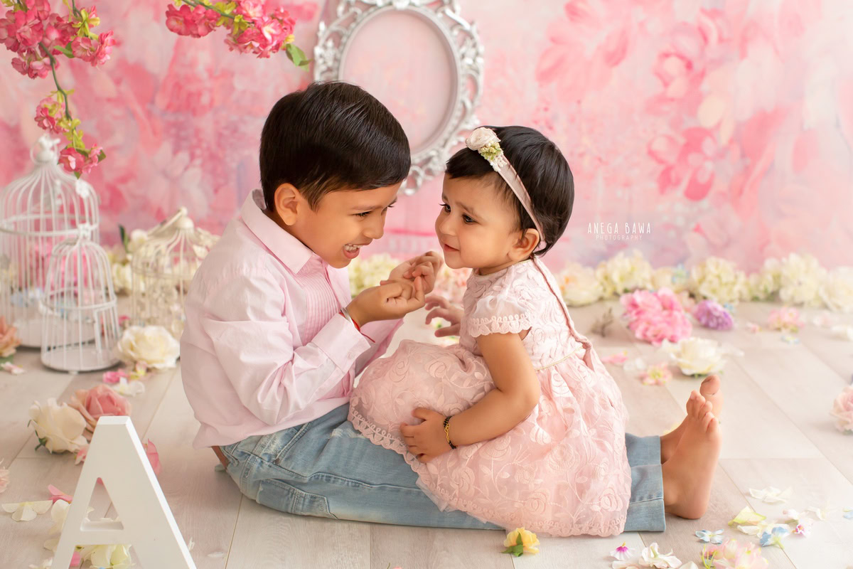 Captured in Gurgaon by Anega Bawa, Delhi, this delightful photo features a 14-month-old girl and her sibling against a soft pink backdrop. A silver frame adorns the wall behind them, while castles and an alphabet frame rest on the floor, adding a whimsical touch to the scene. It's a precious moment shared between siblings, beautifully preserved by the photographer.