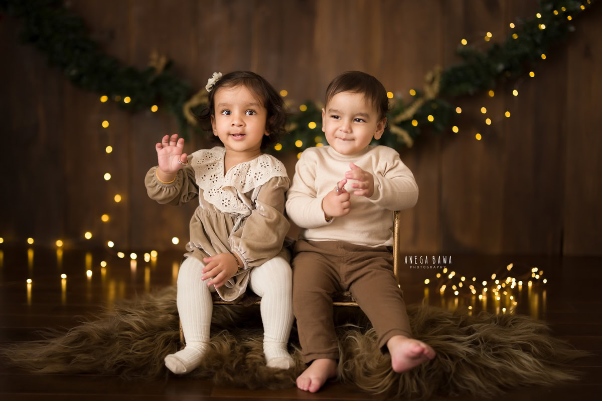 Captured during a sitter photo session by Anega Bawa in Delhi, Gurgaon, this delightful image features two siblings, aged 14 months and 1 year old, seated on a brown rug against a brown backdrop adorned with golden lights.