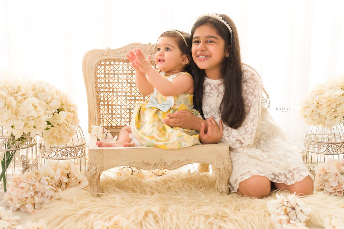 Girl with her sibling seated on a wooden chair atop a beige rug amidst ivory flowers and white castles against a pristine white backdrop, marking their journey from 14 months to 1 year old during their first birthday photoshoot in Delhi by Anega Bawa, Gurgaon, Noida.