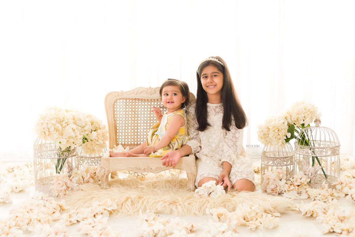 During a baby pre-birthday photoshoot in Delhi, Anega Bawa, Gurgaon, and Noida, two siblings, aged 14 months and 1 year old, sit on a wooden chair adorned with ivory flowers. Behind them, a backdrop of pure white enhances the innocence of their bond, while delicate white castles add a whimsical touch to the scene.