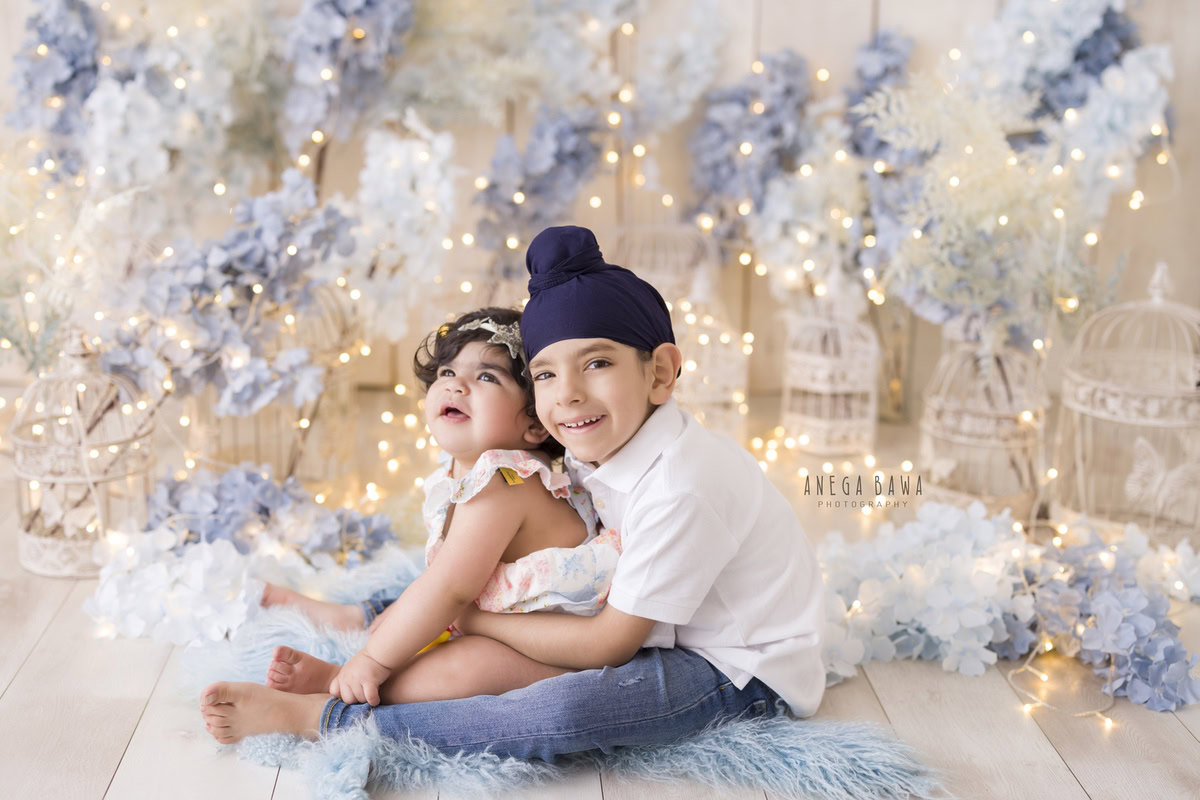 In this captivating toddler photoshoot by Anega Bawa in Delhi, Gurgaon, and Noida, two adorable siblings, aged 14 months and 1 year old, are depicted in a dreamy scene. They are surrounded by white castles and twinkling fairy lights, creating a magical ambiance. The soft blue rug complements the scene perfectly, adding a touch of whimsy to the setting. Together, the siblings explore this enchanting world, their bond evident in their playful interactions. These precious moments captured by the photographer will surely be treasured by the family for years to come.