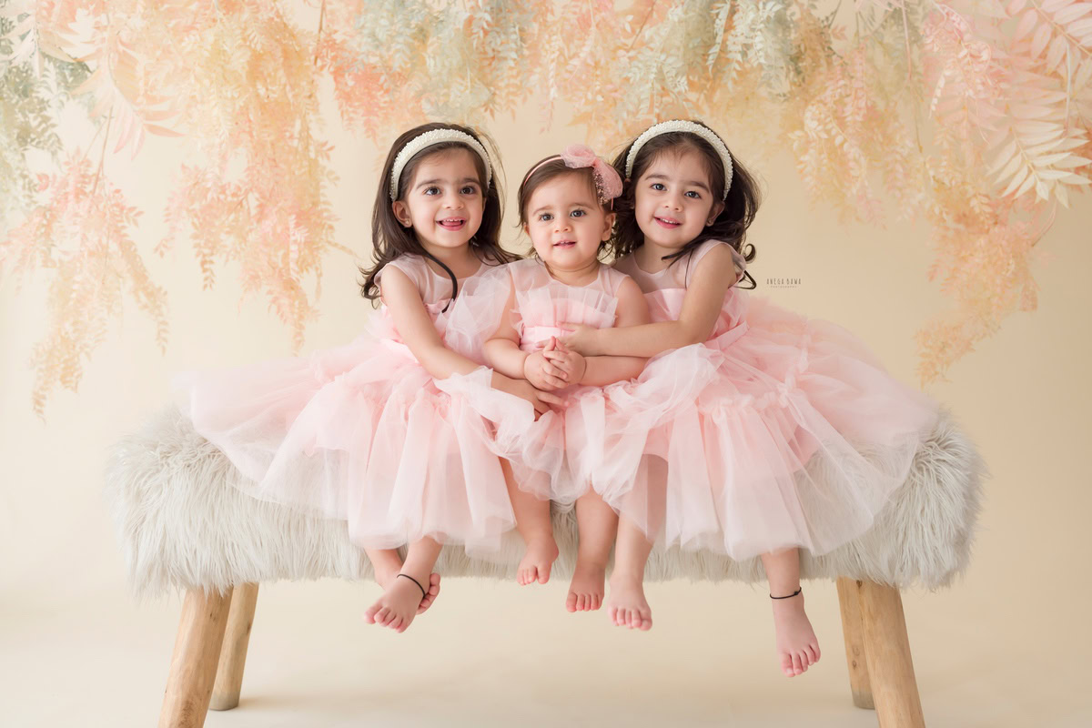 In this heartwarming toddler photoshoot by Anega Bawa in Delhi, Gurgaon, and Noida, two adorable siblings, aged 14 months and 1 year old, share a special moment together. Sitting on a plush white rug and perched on a stool, they are framed against a backdrop of leafy peach, creating a soft and serene atmosphere. Their innocence and connection shine through, capturing the essence of sibling love in a tender and timeless way. These precious moments, skillfully captured by the photographer, will be cherished by the family for years to come.