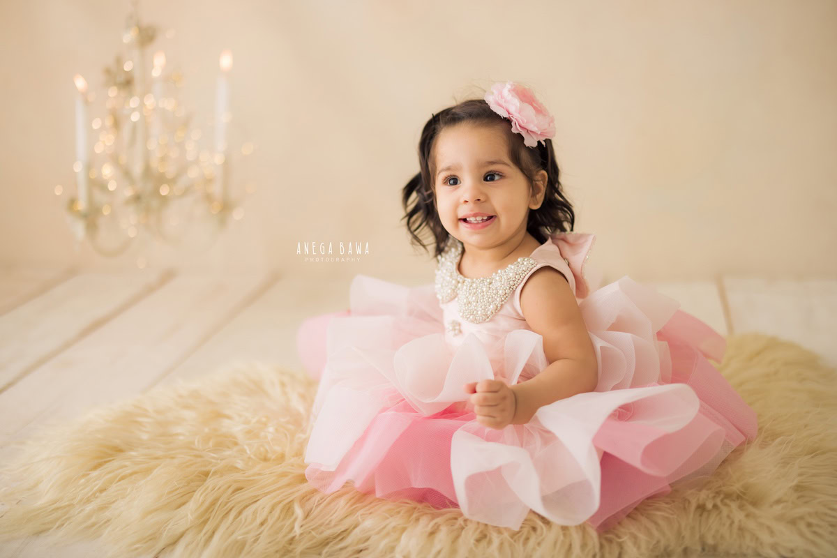 In this enchanting toddler photoshoot by Anega Bawa in Delhi, Gurgaon, and Noida, a charming girl, aged 10 months to 1 year old, sits on a cozy beige rug against a backdrop of soothing beige. The warm glow of golden lights and the flicker of candle stands add a touch of magic to the scene, creating a whimsical ambiance. With her innocent gaze and playful spirit, she captivates the viewer, inviting them into her world of wonder and imagination. Through the lens of the photographer, these precious moments are immortalized, preserving the innocence and joy of childhood for eternity.