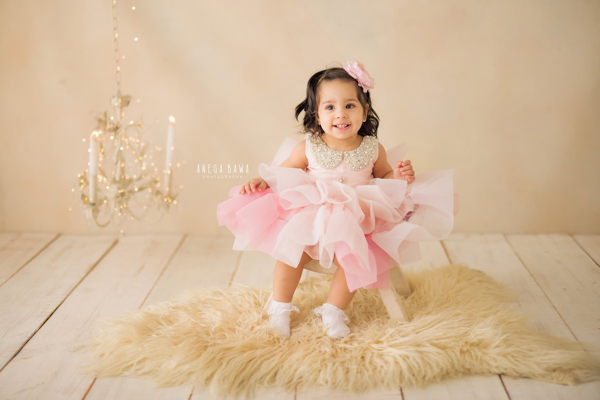 In this delightful toddler photoshoot by Anega Bawa in Delhi, Gurgaon, and Noida, a charming girl, aged 10 months to 1 year old, sits on a soft beige rug, surrounded by the warm glow of golden lights. Against a backdrop of serene beige, the flicker of candle stands adds an enchanting touch to the scene. With her innocent gaze and playful demeanor, she invites viewers into her world of wonder and imagination. Through the lens of the photographer, these precious moments are captured, preserving the joy and innocence of childhood for eternity.