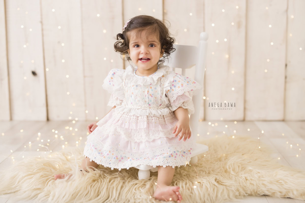 In this charming portrait by Anega Bawa in Gurgaon, Delhi, a 14-month-old girl sits on a beige rug against a serene beige backdrop. The soft glow of fairy lights adds a magical touch to the scene, capturing the innocence and beauty of childhood in a timeless photograph.