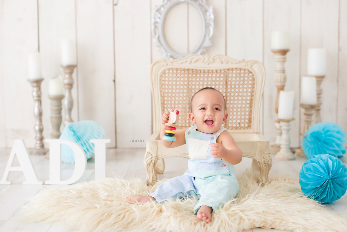 Girl sitting on a cozy beige rug on a wooden chair beside a candle stand and a silver frame on the wall against a serene beige backdrop adorned with pompoms, commemorating her journey from 14 months to 1 year old during her first birthday photoshoot in Delhi by Anega Bawa, Gurgaon, Noida.
