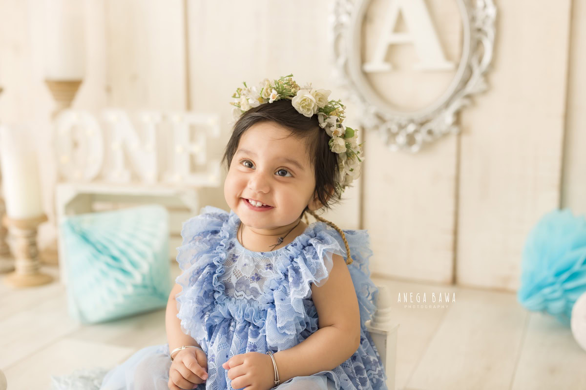 In this endearing toddler photoshoot captured by Anega Bawa in Delhi, Gurgaon, and Noida, a sweet girl, aged 9 months to 1 year old, sits gracefully in a charming blue dress adorned with a tiara band and pompom. Against a soothing beige backdrop, she is surrounded by the warm glow of candle stands and the whimsy of an alphabet frame adorning the wall. With each click of the camera, Anega Bawa beautifully immortalizes these precious moments, capturing the innocence and joy of childhood.