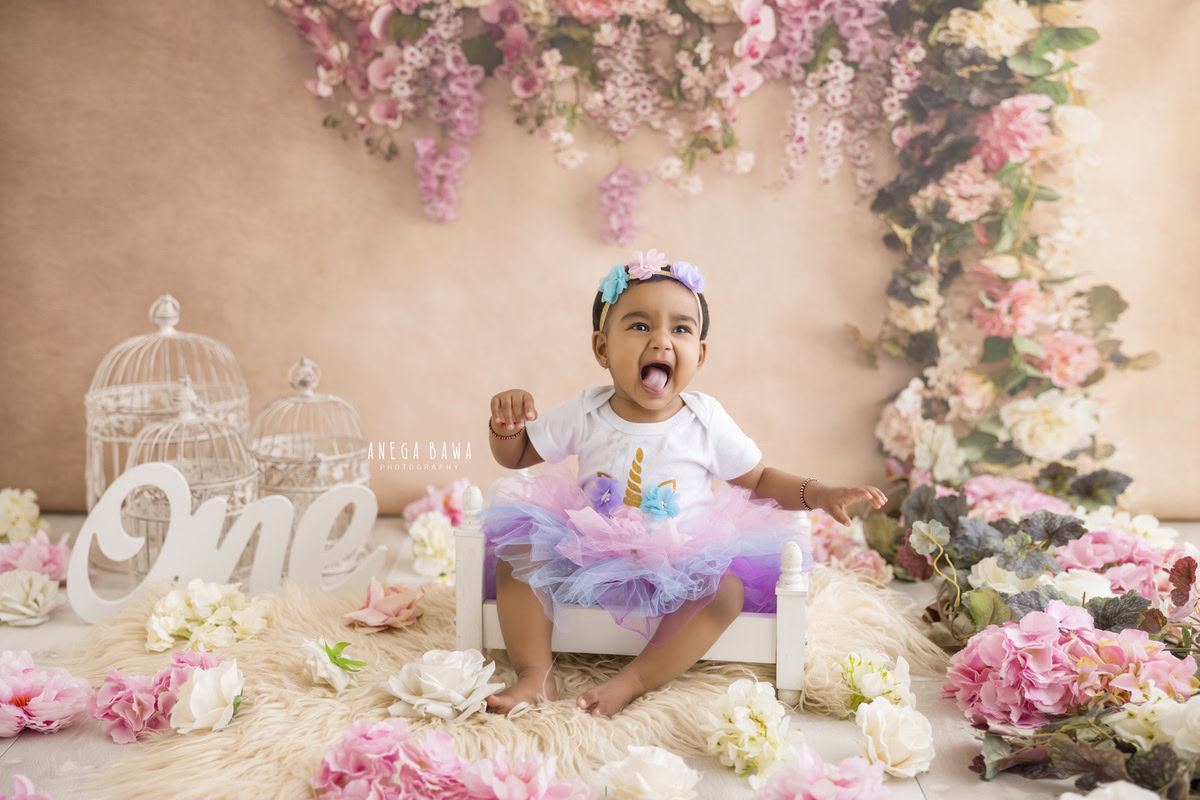 In this delightful toddler photoshoot by Anega Bawa in Delhi, Gurgaon, and Noida, a charming girl, aged 9 months to 1 year old, sits comfortably in a white cot against a serene peach backdrop. Surrounding her are enchanting castles, adding a touch of whimsy to the scene. Adorned with a floral fringe, she exudes innocence and grace, captured beautifully by Anega Bawa's lens. These timeless images serve as cherished memories of this precious stage of childhood.
