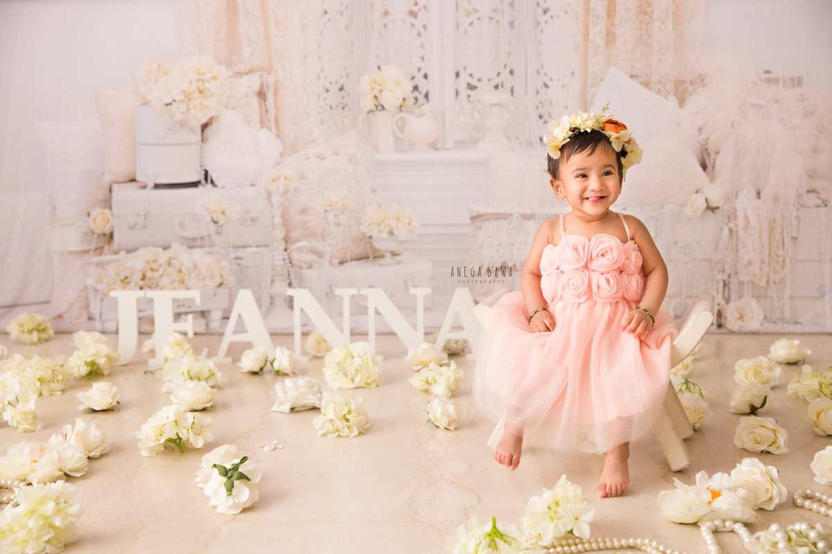 Captured by Anega Bawa in Delhi, this delightful photograph features a 14-month-old girl sitting gracefully on a curvy chair amidst a pristine white setup. Ivory flowers and pearls adorn the floor, adding an elegant touch to the scene. The little girl wears a charming pink dress and a tiara band, exuding a sense of innocence and sweetness. Behind her, white castles complete the enchanting backdrop, creating a magical ambiance that perfectly complements her youthful presence.