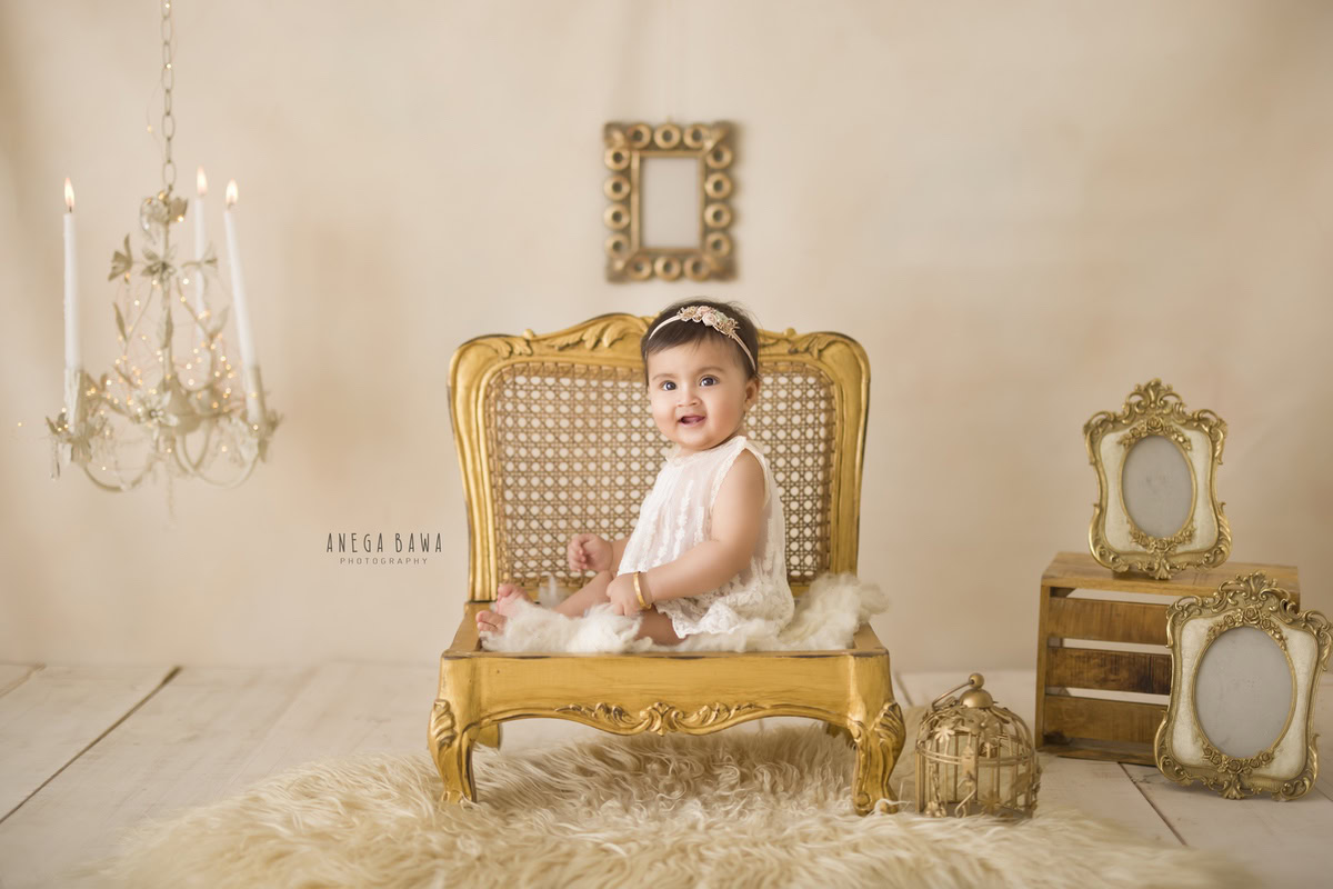 In this delightful portrait captured by Anega Bawa in Gurgaon, a 15-month-old girl sits gracefully on a golden chair against a serene beige backdrop. The scene is adorned with elegant frames on the wall and a delicate candle stand, adding a touch of sophistication to the composition. Anega Bawa's expert photography skillfully captures the innocence and charm of the young sitter, creating a timeless memory to cherish for years to come.