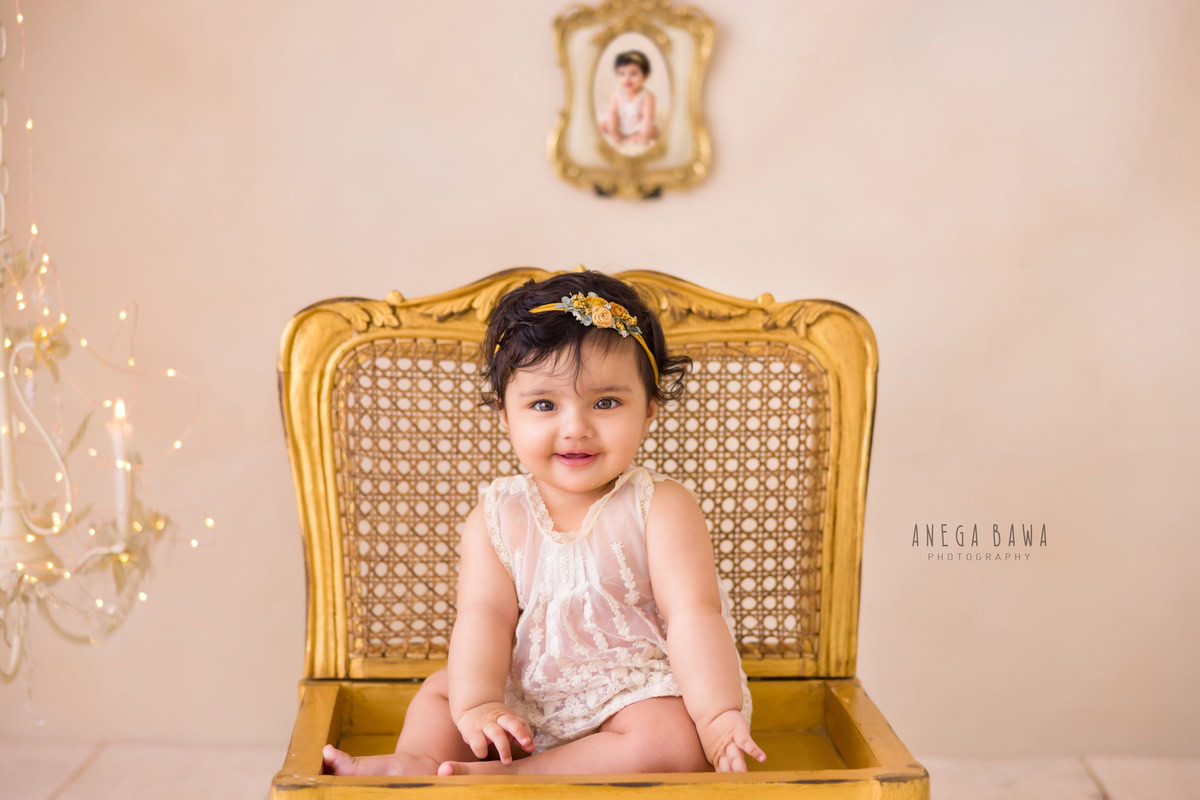 Girl sitting on a regal golden chair beside a golden photoframe on the wall and a candle stand adorned with lights against a serene beige backdrop, commemorating her journey from 15 months to 1 year old during her first birthday photoshoot in Delhi by Anega Bawa, Gurgaon, Noida.