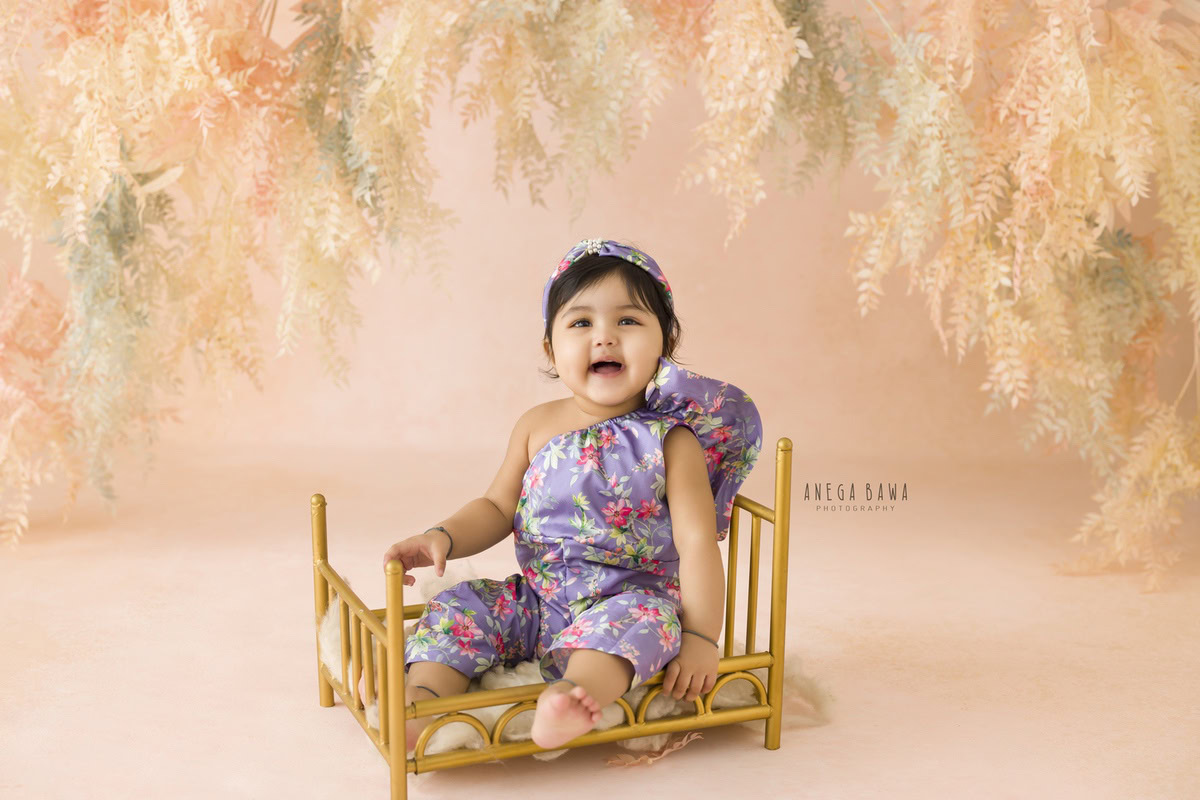 In this delightful toddler photoshoot captured by Anega Bawa in Delhi, Gurgaon, and Noida, a sweet girl, aged 9 months to 1 year old, sits gracefully in a golden cot adorned in a floral jumpsuit. The backdrop, adorned with leafy peachy hues, adds a touch of natural elegance to the scene. Through Anega Bawa's lens, the innocence and charm of childhood are beautifully preserved, creating timeless memories for the family to cherish.