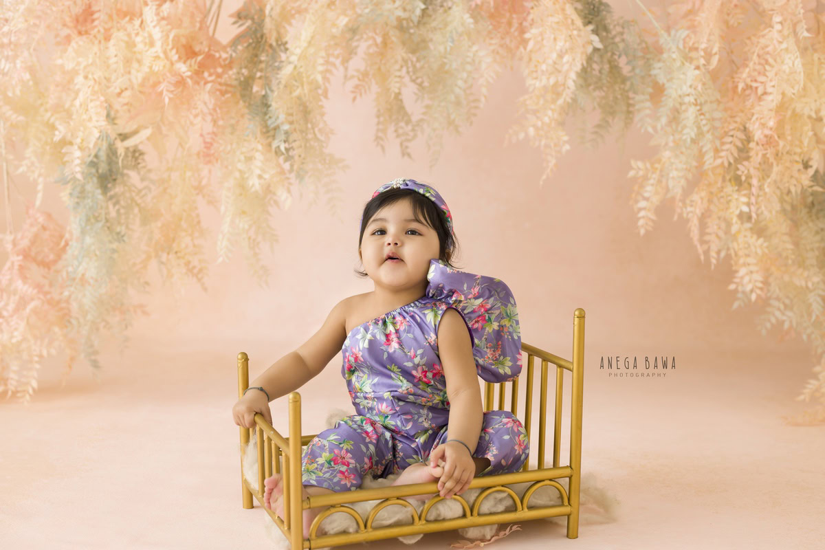 In this enchanting toddler photoshoot captured by Anega Bawa in Delhi, Gurgaon, and Noida, a lovely girl, aged 9 months to 1 year old, sits gracefully in a golden cot against a backdrop adorned with leafy peach hues. Through Anega Bawa's lens, the innocence and charm of childhood are beautifully preserved, creating timeless memories for the family to cherish.