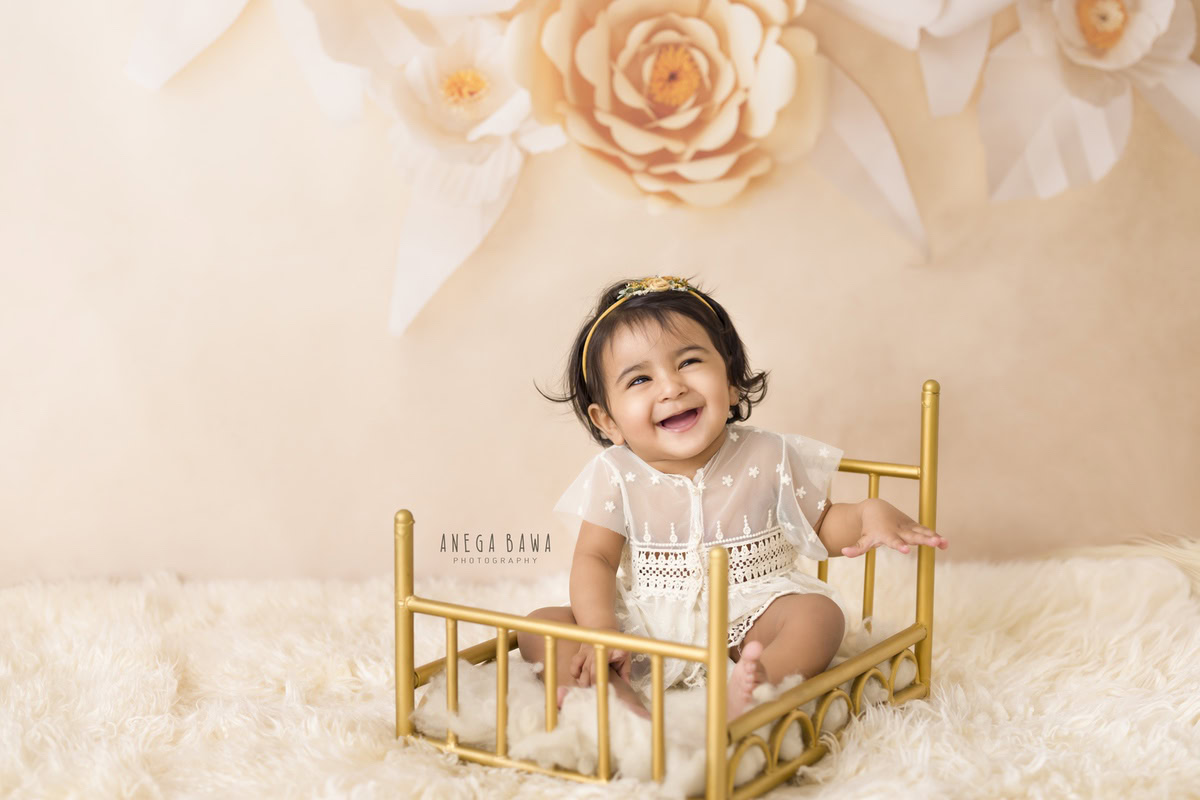 Captured by Anega Bawa in Gurgaon and Delhi, this endearing photograph features a 15-month-old girl seated in a golden cot. Adorned with a soft white rug, the cot is set against a backdrop of delicate peach florals, creating a charming ambiance. The girl's adorable expression, coupled with her cute net top, adds a touch of innocence and sweetness to the portrait. Anega Bawa expertly captures the essence of childhood in this lovely composition.