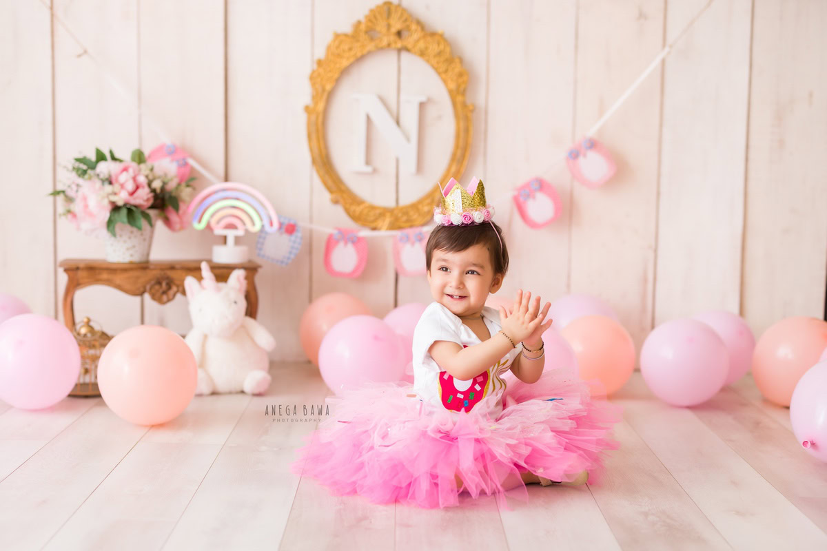 In this charming photograph taken by Anega Bawa in Delhi, Gurgaon, a girl, 15 months old, is captured seated in a pink dress against a beige backdrop. An alphabet frame on the wall and nude-colored balloons add to the whimsical ambiance.