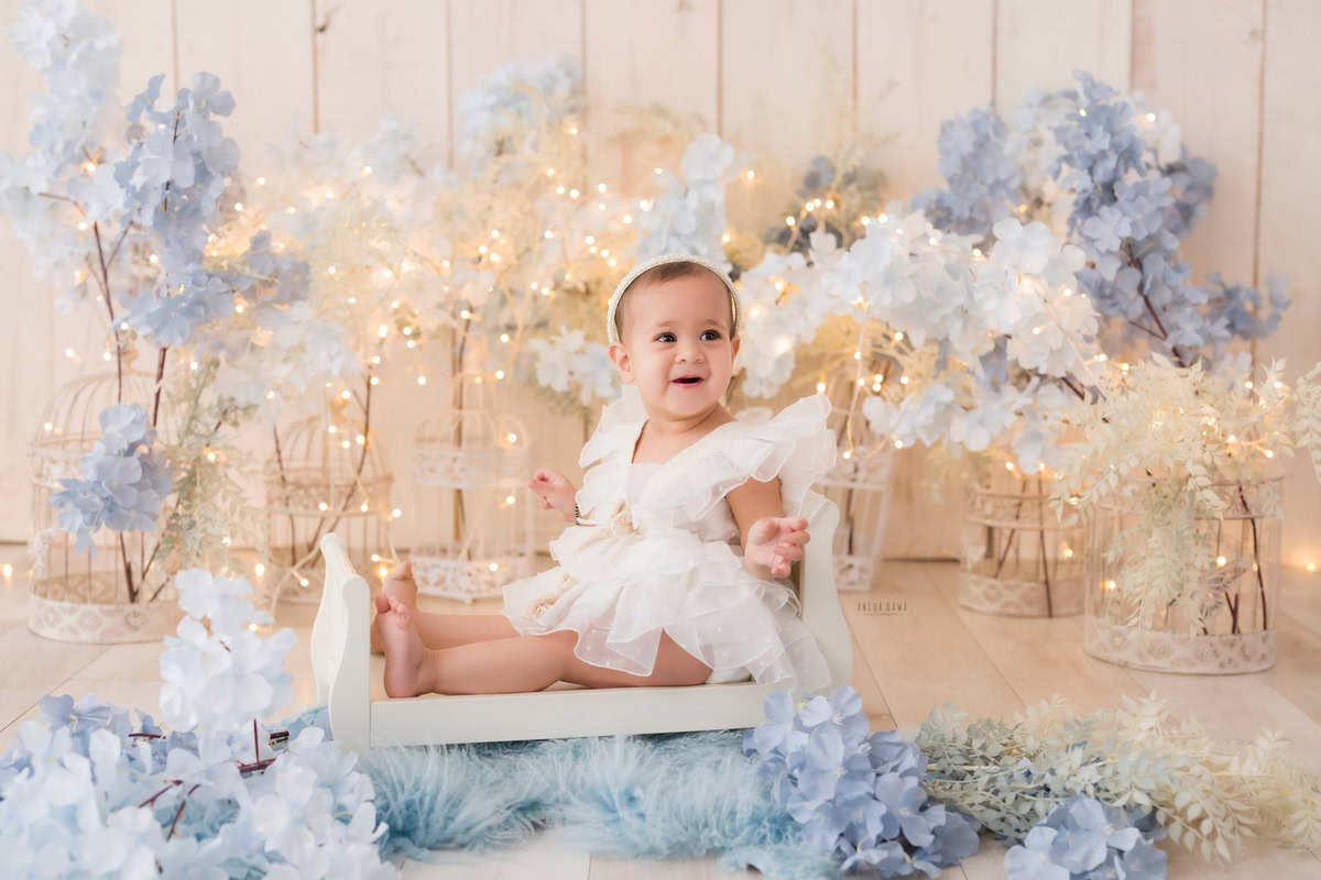 Girl sitting in a pristine white cot on a blue rug surrounded by blue and white flowers and castles against a beige backdrop, with fairy lights illuminating the scene, marking her journey from 15 months to 1 year old during her first birthday photoshoot in Delhi by Anega Bawa, Gurgaon, Noida.