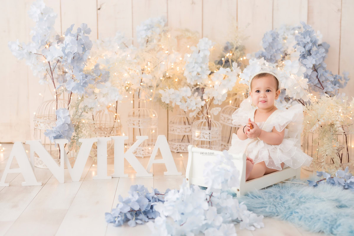For a baby pre-birthday photoshoot in Delhi, Anega Bawa, Gurgaon, and Noida, a 15-month-old girl sits comfortably in a white cot. She is surrounded by a blue rug and delicate white castles. On the floor, a name frame adds a personalized touch to the scene, all against a soothing beige backdrop.