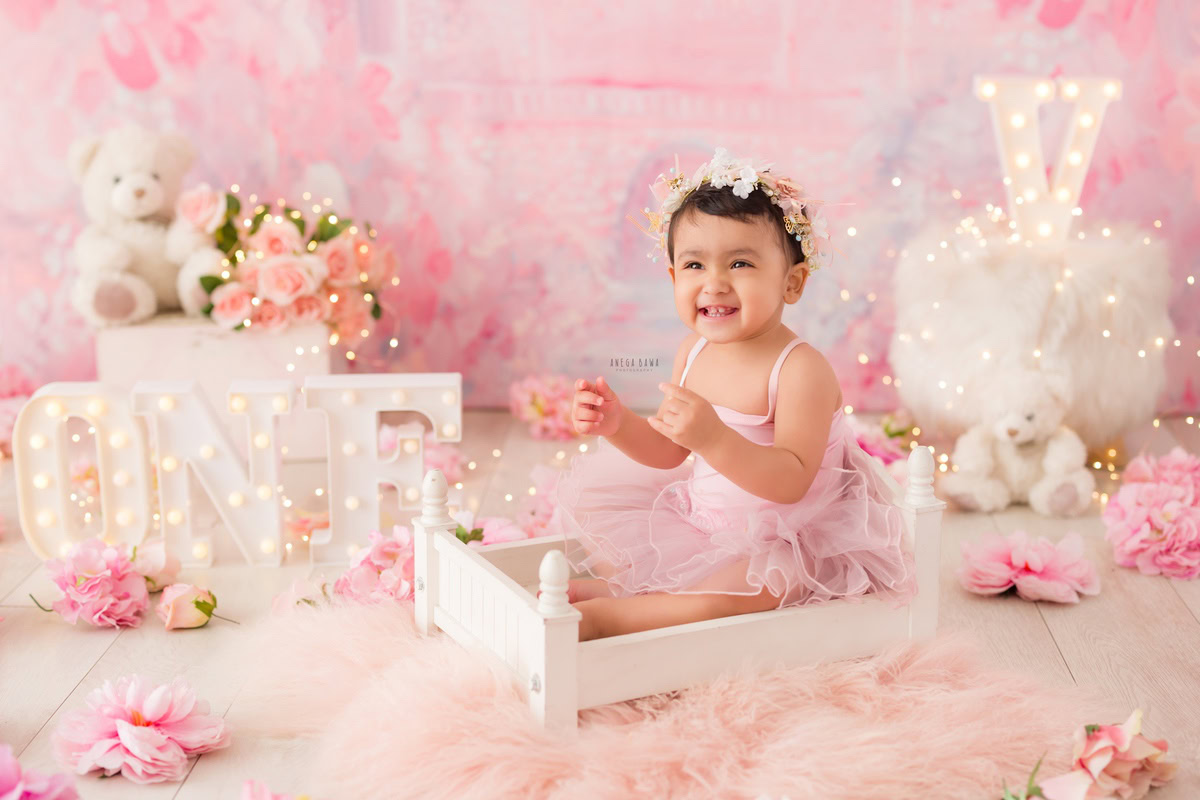 For a baby pre-birthday photoshoot in Delhi, Anega Bawa, Gurgaon, and Noida, a 15-month-old girl sits gracefully in a white cot. The backdrop is a soft pink, complemented by the warm glow of fairy lights. Adorable teddy bears add a playful touch to the scene, while the girl wears a charming tiara band, adding a touch of royalty to the ambiance.