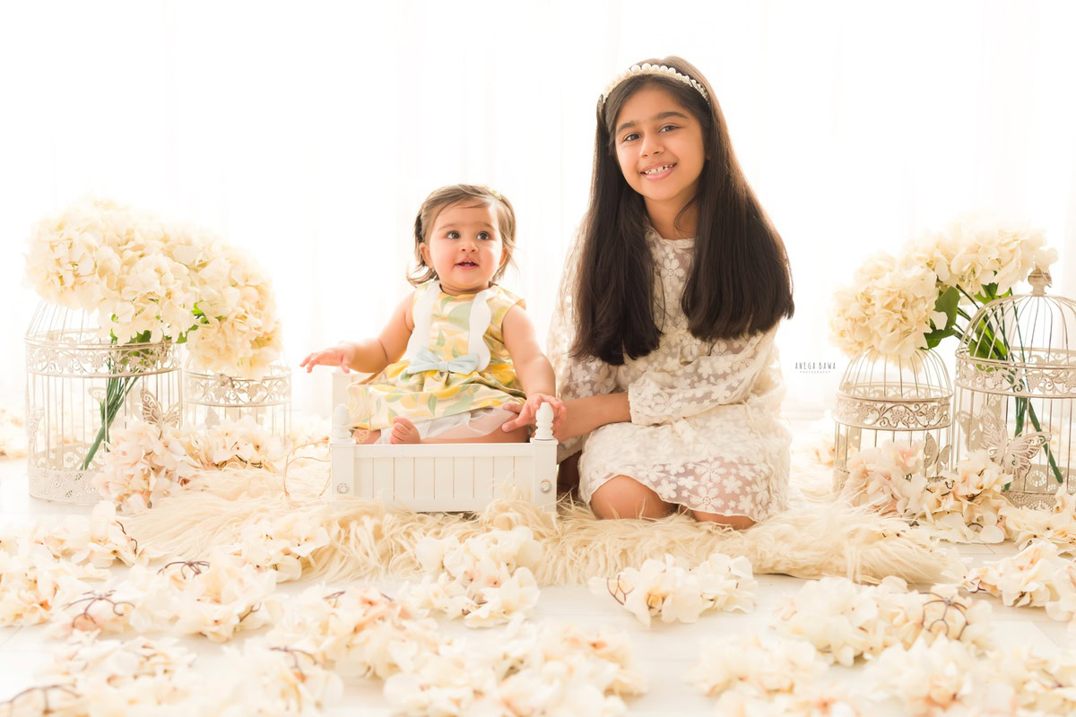 Girl seated in a pristine white cot with her sibling amidst ivory flowers against a pristine white backdrop, accompanied by castles, marking her journey from 15 months to 1 year old during her first birthday photoshoot in Delhi by Anega Bawa, Gurgaon, Noida.