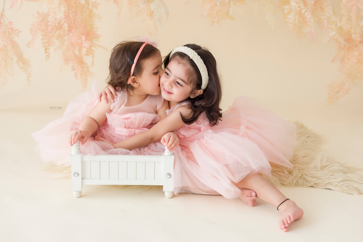 In this heartwarming capture by Anega Bawa, set against a leafy beige backdrop, a precious moment unfolds as a toddler, aged 15 months to 1 year, sits in a white cot alongside a sibling. The tender kiss shared between the siblings encapsulates the pure bond of love and companionship. Through Anega Bawa's lens, this beautiful moment is immortalized, reflecting the joy and warmth of family connections in the timeless art of photography.