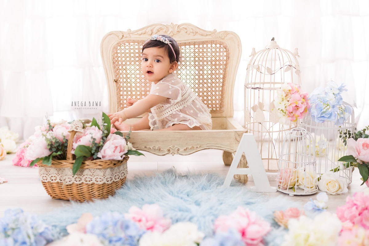 Captured by Anega Bawa in Delhi, Gurgaon, this delightful photo features a 15-month-old girl seated on a beige chair. Against a blue rug and white castles backdrop, she's surrounded by a floral basket with pink and white flowers, adding a touch of charm to the scene.