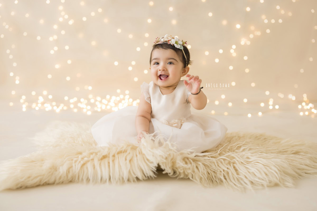 Capture the enchanting innocence of your little one with Anega Bawa Photography in Delhi and Gurgaon. Our sitter photography session features your 15-month to 1-year-old girl seated on a cozy beige rug, surrounded by twinkling fairy lights against a pristine white backdrop. Don't miss the opportunity to preserve these precious moments forever. Book your session now!
