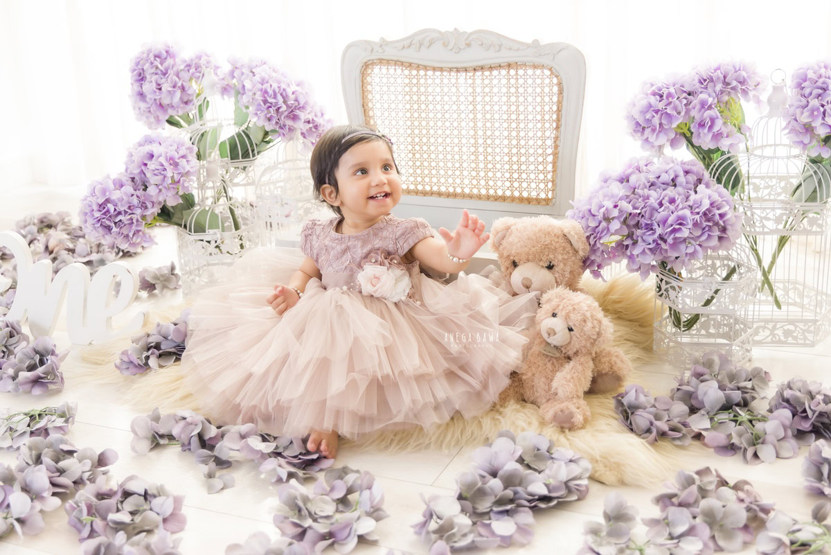 In this delightful scene captured by Anega Bawa, a toddler, aged 8 months to 1 year, sits on a cozy beige rug surrounded by cuddly teddy bears and charming white castles. A bunch of vibrant flowers adds a pop of color to the serene white backdrop. Through Anega Bawa's lens, this enchanting moment is preserved, showcasing the innocence and wonder of childhood in the art of toddler photography.