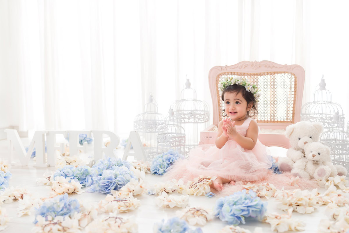 In this endearing portrait by Anega Bawa, a toddler, aged 15 months to 1 year, sits gracefully on the floor surrounded by delicate blue and white flowers. Positioned beside a quaint wooden chair against a pristine white backdrop, the scene exudes a timeless charm. Through Anega Bawa's lens, the innocence and beauty of childhood are artfully captured in this toddler photoshoot, serving as a cherished memento for years to come.