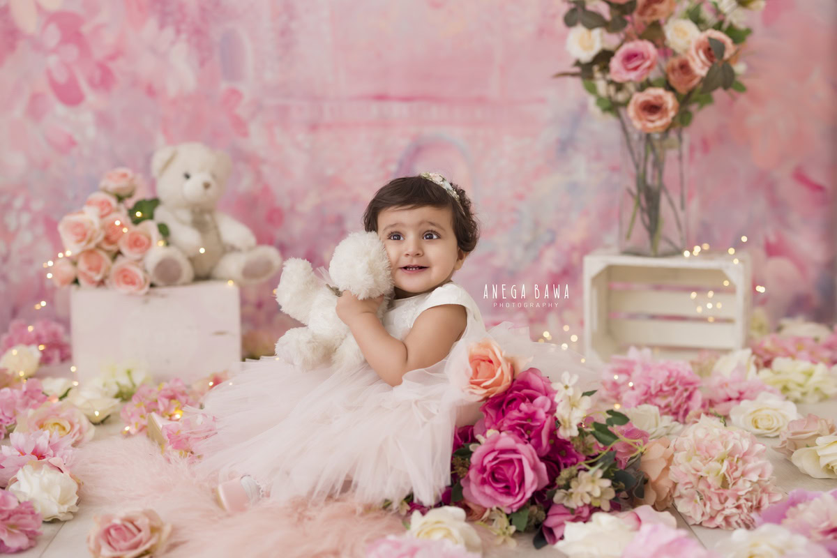 In this charming portrait by Anega Bawa, captured in Delhi and Gurgaon, a 15-month-old girl sits on the floor, clutching a teddy bear. Against a lovely pink backdrop adorned with delicate pink flowers, the innocence and sweetness of childhood are beautifully highlighted. Anega Bawa's expertise as a photographer shines through in this captivating image, preserving a precious moment in time for the young sitter and her family.