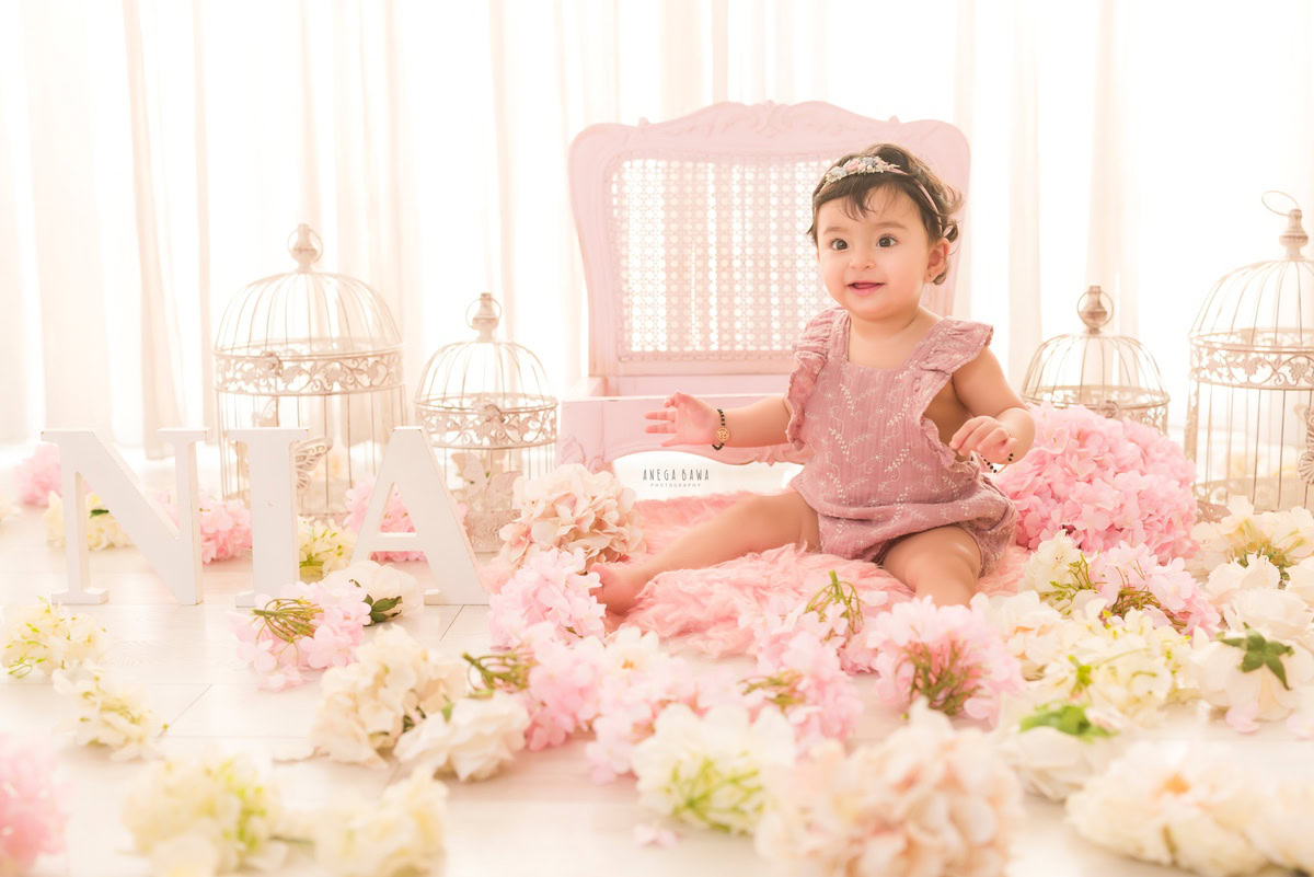 For a baby pre-birthday photoshoot in Delhi, Anega Bawa, Gurgaon, and Noida, a 15-month-old girl sits amidst a scene of whimsy and charm. She is seated on the floor surrounded by white castles and delicate pink and white flowers. A name frame rests on the floor nearby, adding a personalized touch to the setup. The backdrop is a serene white, allowing the scene to shine with its own enchanting beauty.