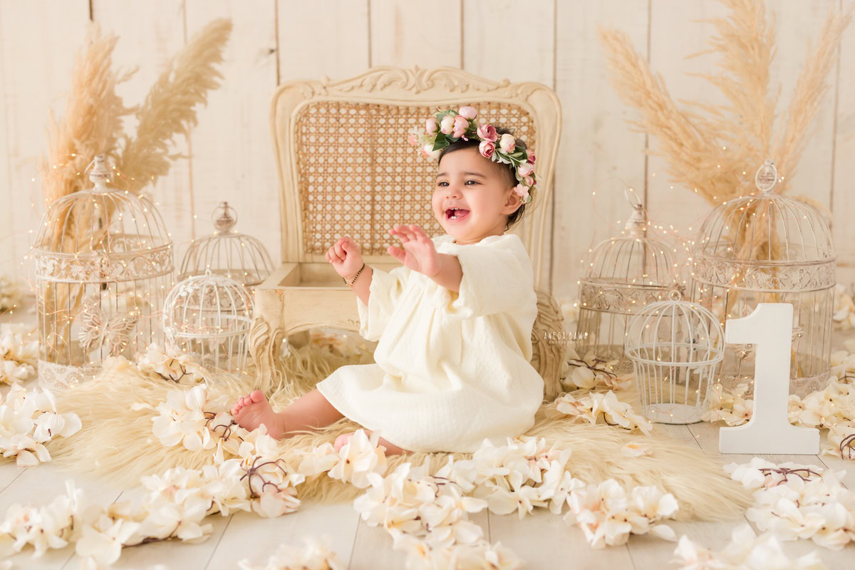 For a baby pre-birthday photoshoot in Delhi, Anega Bawa, Gurgaon, and Noida, a 15-month-old girl is captured in a picturesque scene. She sits on the floor, adorned with a wooden chair, surrounded by delicate white flowers and bushes. White castles add a touch of whimsy to the setting, creating a magical ambiance. The backdrop complements the scene with its serene and enchanting vibe.