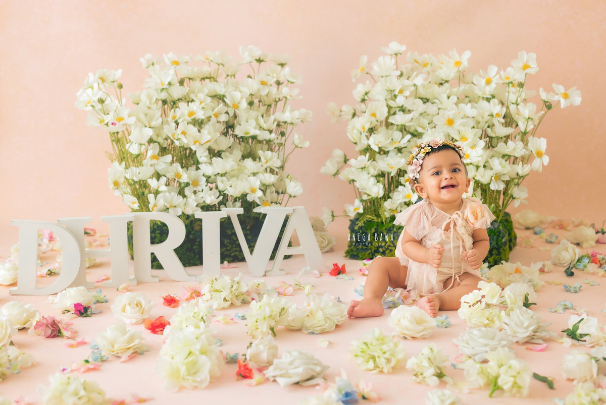 In this enchanting capture by Anega Bawa, a little girl, aged 7 months to 1 year, sits serenely amidst a bed of vibrant floral blooms scattered on the floor, set against a lovely peach backdrop. Adorning the wall behind her is a charming name frame, adding a personalized touch to the scene. The soft, natural light enhances the warmth of the image, while the delicate white flowers accentuate the innocence and beauty of the moment. Through Anega Bawa's lens, this snapshot immortalizes the joy and wonder of early childhood, creating a cherished memory for years to come.