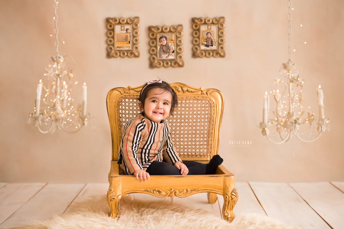 In this enchanting photo shoot by Anega Bawa in Delhi, Gurgaon, a 15-month-old girl is captured sitting on a golden chair against a beige backdrop. Adorned in a Burberry top, she exudes elegance and style. The scene is enhanced by photo frames on the wall, golden lights, a candle stand, and a beige rug, creating a warm and inviting atmosphere.