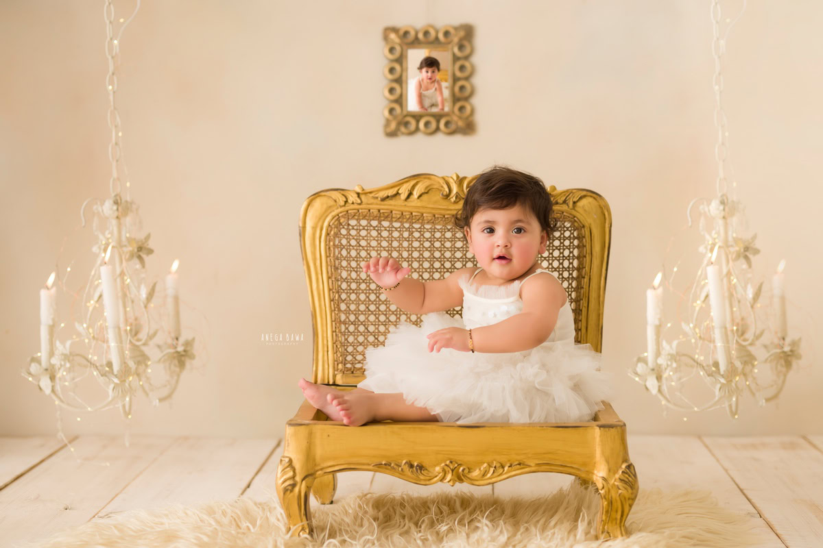 For a baby pre-birthday photoshoot in Delhi, Anega Bawa, Gurgaon, and Noida, a 15-month-old girl sits gracefully on a golden chair. Beside her, a candle stand illuminates the scene, casting a warm glow. A photo frame adorns the wall behind her, adding a personal touch to the setup. The beige backdrop provides a neutral and elegant background, allowing the focus to remain on the charming subject.