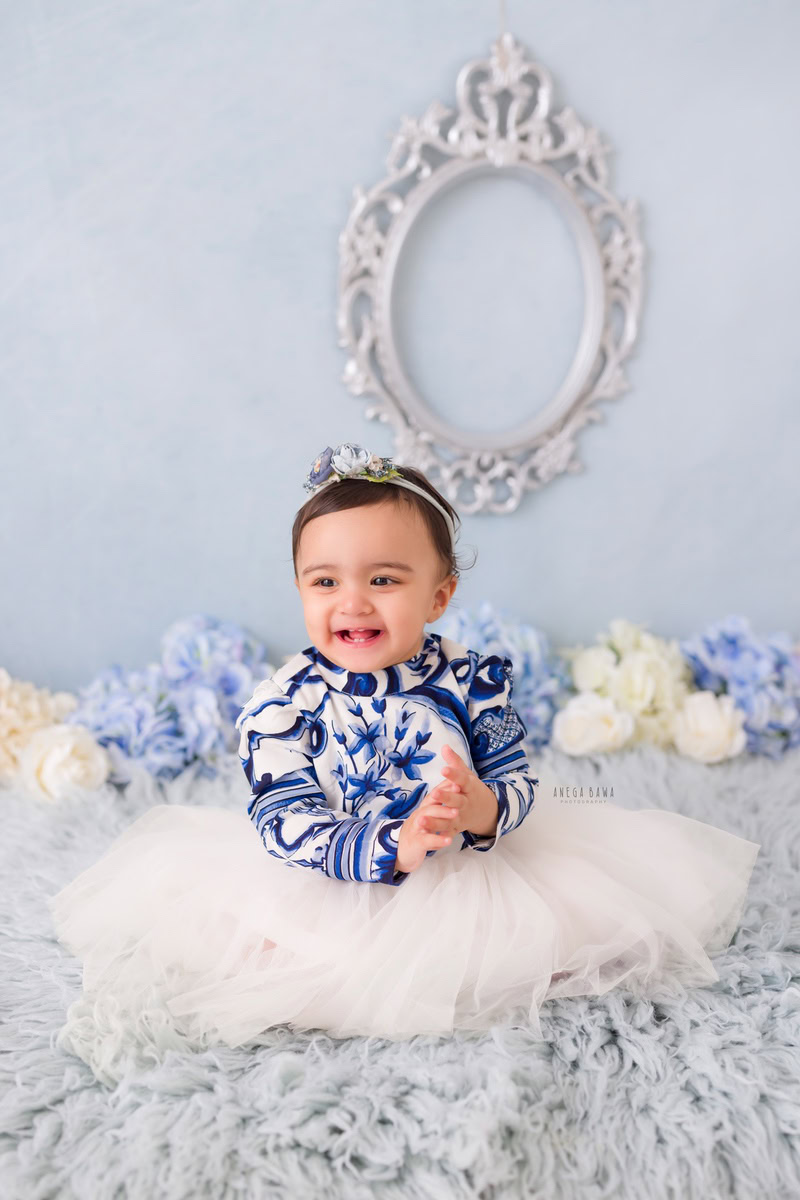 Embrace the ethereal charm of your little princess seated on a soft grey rug, surrounded by delicate blue and white flowers against a serene blue backdrop. With a touch of elegance from a silver frame adorning the wall, Anega Bawa Photography in Delhi and Gurgaon ensures every precious moment is immortalized. Book your sitter photography session now!