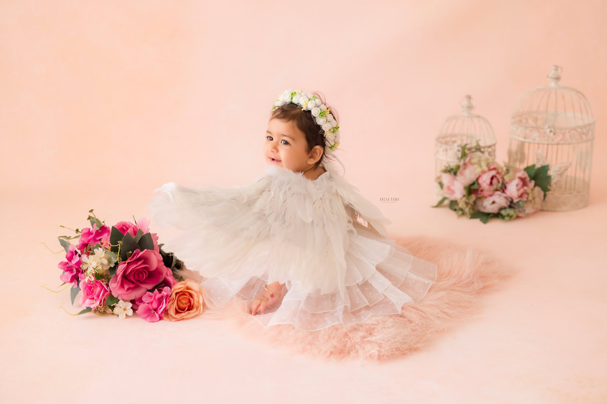 Capture the innocence and grace of your little one as she sits on a peach floor, adorned with a charming tiara band, amidst white castles and a delightful bunch of flowers. Against the backdrop of a soft peach hue, Anega Bawa Photography in Delhi and Gurgaon brings out the purity of childhood in every frame. Book your sitter photography session today and treasure these precious moments forever!