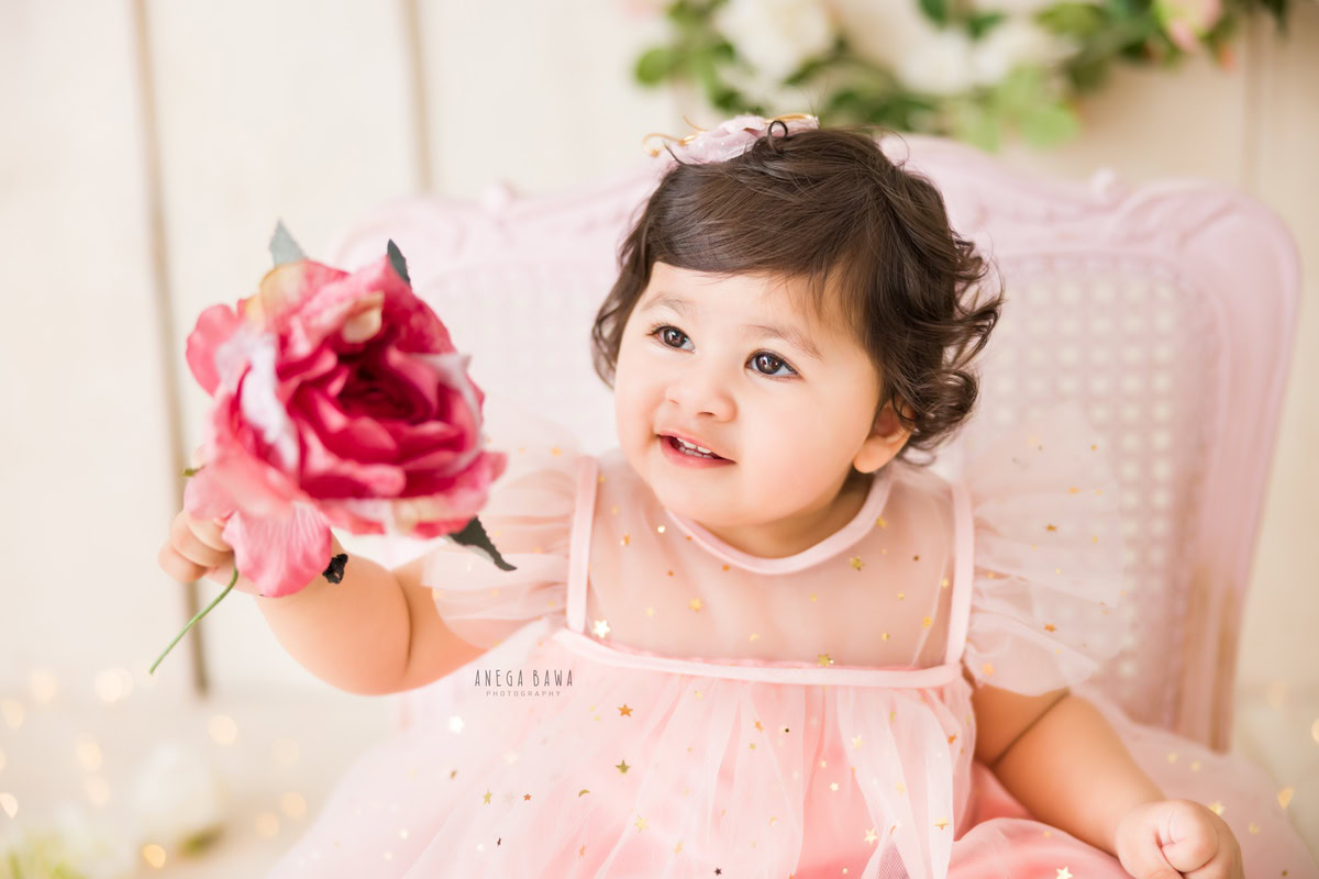In this endearing photograph taken by Anega Bawa in Delhi and Gurgaon, a 15-month-old girl sits gracefully on a pink chair, holding a delicate rose. Dressed in a charming pink outfit, she exudes an aura of innocence and beauty against the soft beige backdrop. Anega Bawa's skillful photography captures the essence of childhood in this precious moment, creating a timeless keepsake for the family to cherish.