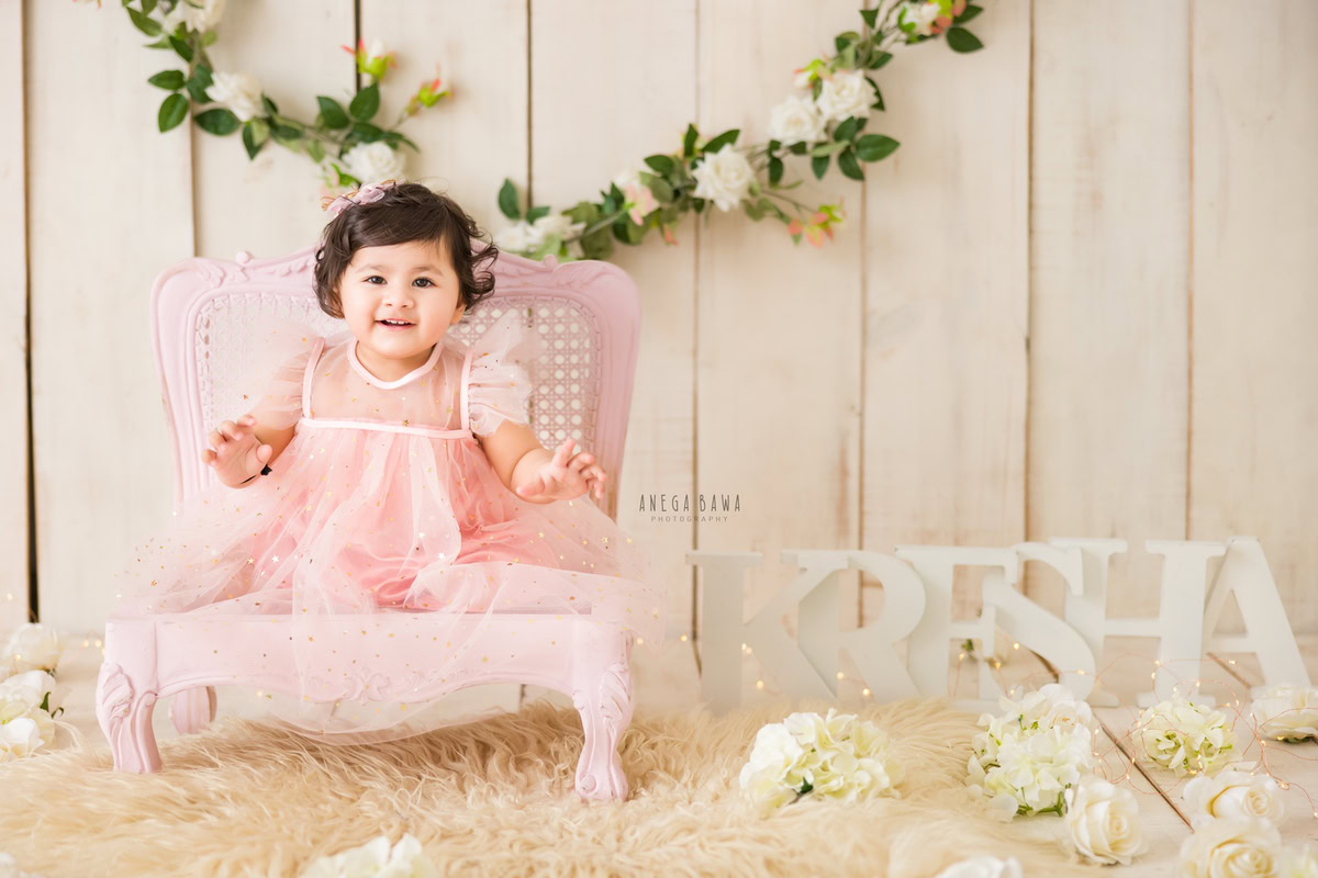 Captured by Anega Bawa in Gurgaon and Delhi, this delightful image features a 16-month-old girl seated on a pink chair, adorned in a lovely pink dress. Against the backdrop of a beige rug and surrounded by delicate white flowers, the young sitter emanates an aura of innocence and charm. Through Anega Bawa's lens, the beauty of childhood is immortalized in this enchanting moment, creating a precious memory for the family to treasure.