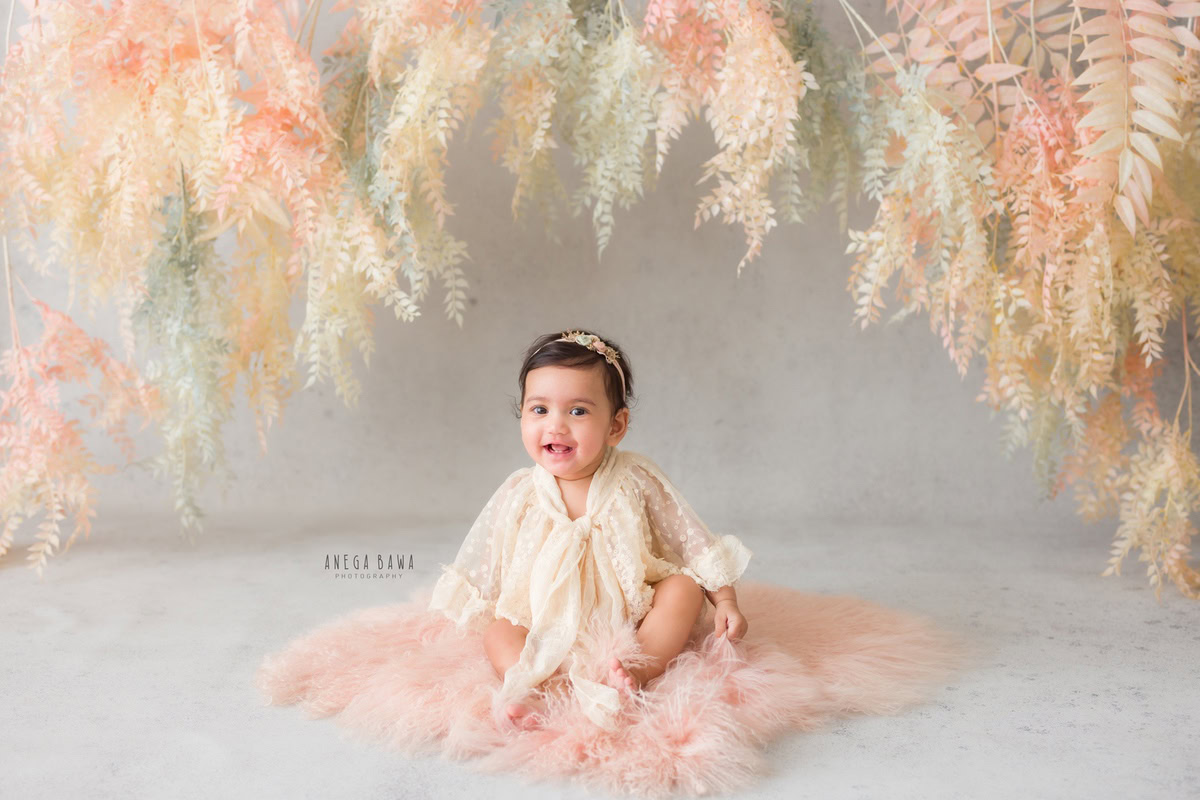 Captured by the talented lens of Anega Bawa, this delightful image features a precious moment of a girl aged 7 months to 1 year. Seated gracefully on a soft pink rug, she exudes innocence and charm against a striking blue backdrop adorned with lush green foliage. Anega Bawa skillfully captures the juxtaposition of the girl's delicate presence against the vibrant backdrop, creating a visually captivating composition. This enchanting toddler photoshoot, set in Delhi, Gurgaon, or Noida, showcases the magic of childhood in every frame.