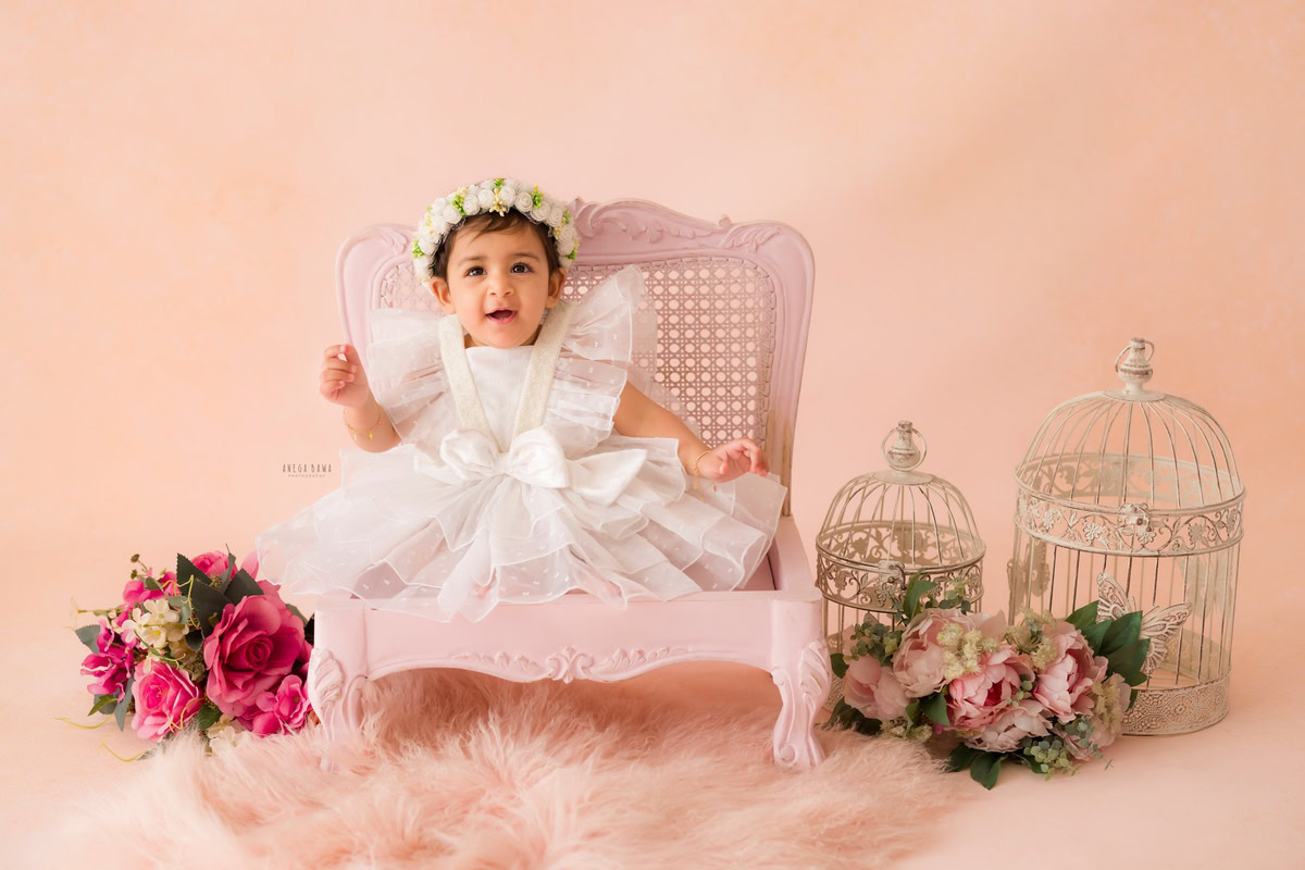 Girl seated on a charming pink wooden chair atop a pink rug surrounded by white castles and a bunch of flowers against a delightful peach backdrop, marking her journey from 16 months to 1 year old during her first birthday photoshoot in Delhi by Anega Bawa, Gurgaon, Noida.