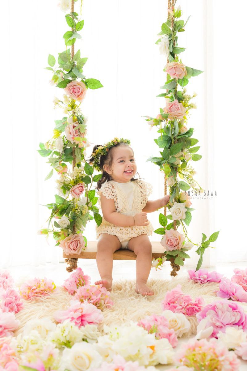 In this endearing capture by Anega Bawa, a girl aged 7 months to 1 year sits gracefully on a swing adorned with delicate pink and white flowers. Adding to her enchanting charm is a cute tiara band adorning her head. Against the backdrop of the bustling cityscape of Delhi, Gurgaon, or Noida, Anega Bawa expertly captures the innocence and joy of childhood. This toddler photoshoot is a testament to the magic of fleeting moments, beautifully preserved in each frame.