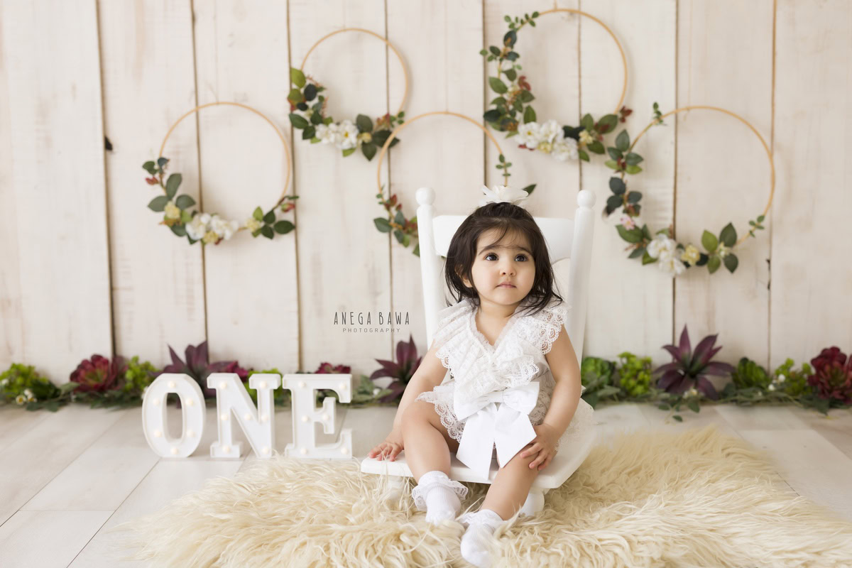 In this captivating shot from Anega Bawa's lens, a girl, aged 7 months to 1 year, sits elegantly on a white chair, surrounded by the warmth of a beige rug. The wooden floral frames adorning the wall add a touch of rustic charm to the scene, while the beige backdrop provides a serene backdrop for the toddler's playful innocence. Anega Bawa's skilled photography captures the essence of childhood in this delightful toddler photoshoot, set against the vibrant backdrop of Delhi, Gurgaon, or Noida.