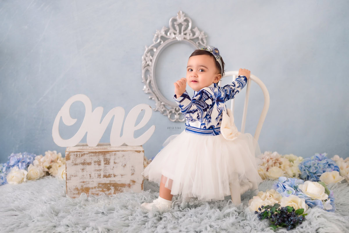 Adorned in innocence, the little girl sits atop a white chair, her presence gracing a serene setting accentuated by a grey rug and delicate blue and white flowers. At 16 months old, she embodies the essence of childhood, captured beautifully by Anega Bawa Photography in Delhi and Gurgaon. Don't let these fleeting moments pass by; seize the opportunity to preserve them forever with a sitter photography session.