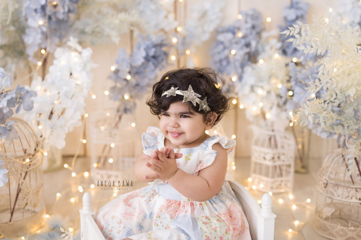 Perched on a white cot against a beige backdrop, the 16-month-old girl is surrounded by an enchanting scene. Delicate blue and white flowers adorn the setting, while miniature white castles add a touch of whimsy. Fairy lights dance around her, casting a magical glow. Anega Bawa captures the ethereal beauty of childhood in this serene moment, offering a timeless portrait of innocence. Capture your little one's precious milestones with sitter photography in Delhi and Gurgaon.