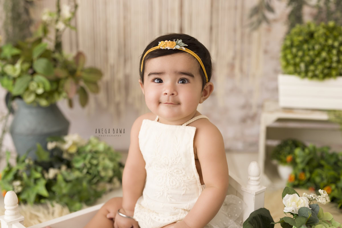 In this enchanting portrait by Anega Bawa, a 13-month-old girl sits gracefully on a white cot adorned with blue pots filled with lush green leaves. Her cute headband adds a touch of innocence to the scene, capturing the essence of childhood. Against the backdrop of Delhi, Gurgaon, or Noida, Anega Bawa's expert photography skillfully preserves this precious moment, immortalizing the beauty of infancy in a timeless image.