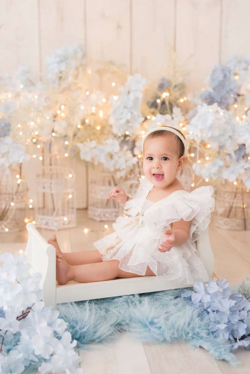 Girl sitting in a pristine white cot surrounded by white castles on a blue rug against a serene beige backdrop, with blue flowers accentuating the scene, marking her journey from 16 months to 1 year old during her first birthday photoshoot in Delhi by Anega Bawa, Gurgaon, Noida.
