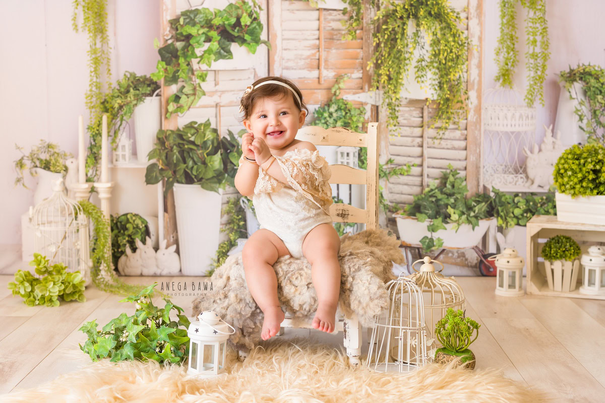 Girl seated on a wooden chair atop a cozy beige rug, surrounded by white pots adorned with green leaves and castles against a serene beige backdrop, marking her journey from 16 months to 1 year old during her first birthday photoshoot in Delhi by Anega Bawa, Gurgaon, Noida.