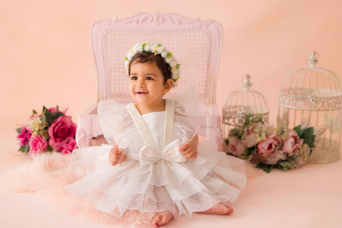 Perched on a pink wooden chair against a soft peach backdrop, the 16-month-old girl exudes charm and sweetness. Delicate white castles complement the scene, adding a touch of whimsy and fairy-tale magic. A bunch of flowers completes the enchanting setting, enhancing the innocence and beauty of childhood captured by Anega Bawa's lens. For timeless portraits that celebrate your little one's milestones, consider sitter photography in Delhi and Gurgaon.